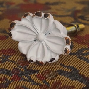 Könnte beinhalten: Ein weißer Möbelknopf aus Metall in Form einer Blume mit mehreren Blütenblättern. Die Ränder der Blütenblätter weisen ein abgenutztes Finish auf, bei dem braune Flecken durch die weiße Farbe sichtbar sind. Ein Metallbolzen ragt aus der Rückseite.