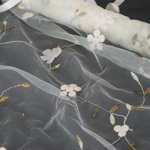 Könnte beinhalten: Weißer Tüllstoff mit gestickten floralen Details. Der zarte Schleier zeigt weiße Blüten und goldfarbene Blätter, mit einem gerollten Abschnitt im Hintergrund. Oben ist ein weißes Band befestigt.