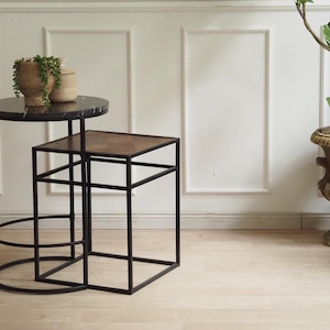Peut inclure: Deux tables d'appoint gigognes avec des cadres en métal noir. La plus grande table a un plateau rond en marbre noir, la plus petite un plateau carré en imitation bois. Une plante décorative dans un pot en pierre est posée sur la plus grande table.