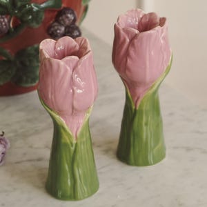 Op de afbeelding: Twee keramische vazen in de vorm van een tulp. De vazen zijn beschilderd met een lichtroze bloem en een groene stengel. De vazen staan op een wit oppervlak. Op de achtergrond staat een rode keramische container met een braambessenmotief.