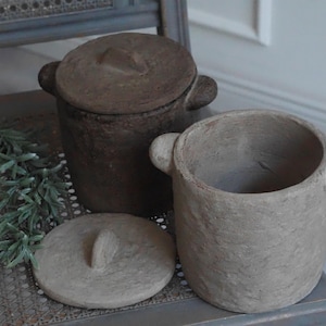 Può includere: Due vasi cilindrici in ceramica con manici laterali e coperchi abbinati. Un vaso è marrone scuro con una superficie ruvida, mentre l'altro è di un colore beige più chiaro e tenue con una finitura opaca e irregolare. Entrambi hanno coperchi.