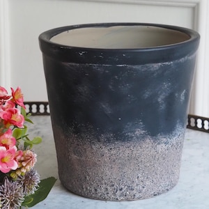 Könnte beinhalten: Zylindrischer, zweifarbiger Blumentopf mit dunkelblauem Oberteil und strukturiertem, hellbraunem Unterteil. Der Innenraum ist hellbeige. Der Topf ist leer. Ein Blumenarrangement mit rosa Blüten befindet sich im Hintergrund.