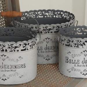 Peut inclure: Trois jardinières ovales en métal avec une finition blanche vieillie et des bords en filigrane gris foncé. Chaque jardinière porte le texte imprimé en noir Belle Jardinière 2, Rue du Pont-Neuf. La plus grande possède une anse en fil de fer.