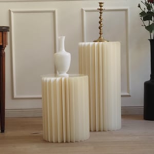 Könnte beinhalten: Zwei cremefarbene, plissierte Papier-Beistelltische in verschiedenen Höhen. Auf dem kleineren Tisch steht eine weiße Vase. Auf dem größeren Tisch steht ein goldener Kerzenhalter. Rechts steht eine schwarze Vase mit Grün und roten Beeren.
