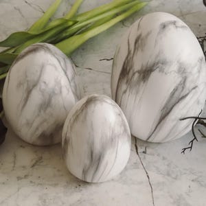 Könnte beinhalten: Drei dekorative, eiförmige Objekte mit weißem und grauem Marmormuster. Die Eier sind unterschiedlich groß, das größte ist etwa 15 cm hoch. Sie sind auf einer Marmoroberfläche mit grünen Blättern und schwarzen floralen Akzenten angeordnet.