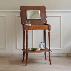 Könnte beinhalten: Ein antiker Holz-Waschtisch mit einem klappbaren Spiegel, einer kleinen Schublade und einem unteren Regal. Der Tisch hat schlanke, gedrechselte Beine und eine warme, natürliche Holzoberfläche. Zwei kleine Glasflaschen stehen auf der Tischplatte.