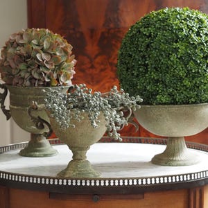 Puede incluir: Tres urnas decorativas con plantas artificiales sobre una mesa redonda. Las urnas son de color verde desgastado y tienen asas ornamentadas. Una urna contiene una esfera verde de boj artificial, otra bayas grises y la tercera flores artificiales.
