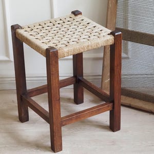 Pode incluir: Um pequeno banco quadrado de madeira com um assento tecido. A estrutura é feita de madeira castanha escura, e o assento é tecido com um material bege claro. O banco tem quatro pernas resistentes e um design simples e clássico.
