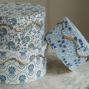 May include: Three decorative hat boxes with floral patterns in shades of blue and white. The boxes are round and rectangular, with gold trim. The boxes are stacked and arranged on a marble surface.
