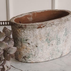 Op de afbeelding: Ovale, verweerde terracotta plantenbak met een verweerde, gebroken witte afwerking. De binnenkant toont de natuurlijke roodbruine klei. De buitenkant heeft hints van groen en bruin, wat op ouderdom duidt. Een decoratief stuk voor planten.