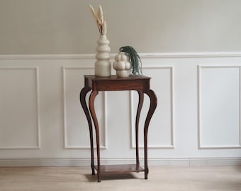 May include: A dark brown, antique-style side table with curved legs and a lower shelf. Two white ceramic vases with decorative plants sit on top. One vase is tall and bulbous, the other is round. The background is a neutral wall.