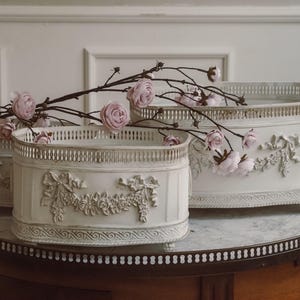 Könnte beinhalten: Drei ovale, weiße Pflanzgefäße mit aufwendigen floralen Reliefdetails und einem dekorativen Rand. Die Pflanzgefäße sind auf einem Tisch mit Marmorplatte angeordnet und mit rosa Blumen und Zweigen geschmückt. Das größte ist etwa 61 cm lang.