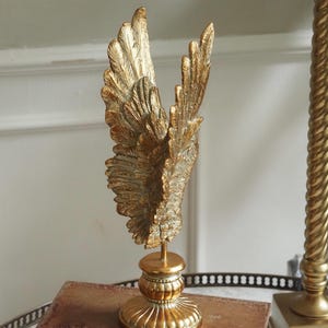 Könnte beinhalten: Eine goldfarbene dekorative Skulptur von Engelsflügeln mit detaillierten Federstrukturen. Die Flügel sind auf einem Sockel montiert. Im Hintergrund ist eine goldfarbene Lampe zu sehen.