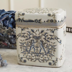 Può includere: Scatola quadrata in ceramica con coperchio, decorata con un dettagliato motivo floreale e di cherubini in blu e oro su sfondo bianco. La scatola ha un bordo dorato e un bordo decorativo sul coperchio.