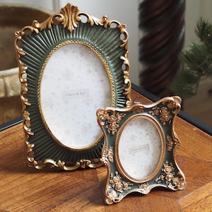 Op de afbeelding: Twee sierlijke fotolijsten in donkergroen en goud. De grotere lijst is ovaal, de kleinere vierkant met bloemendetails. Beide lijsten hebben de tekst "Clayre & Eef". Ze staan op een houten oppervlak.