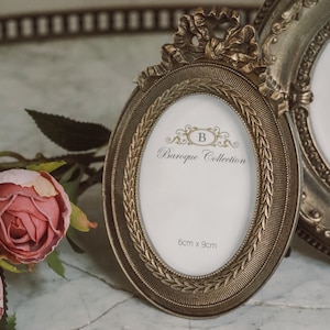 Pode incluir: Moldura oval dourada com a inscrição "Baroque Collection" e dimensões de 6cm x 9cm. A moldura tem um laço decorativo no topo e uma borda detalhada. Uma segunda moldura é visível no fundo.