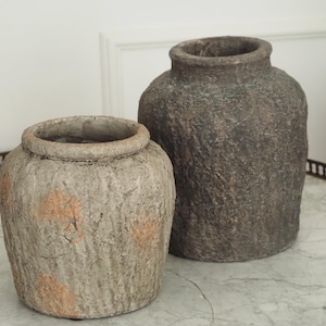 Peut inclure: Deux vases cylindriques aux surfaces texturées et patinées. Le plus petit vase est gris clair avec des taches orange, tandis que le plus grand est brun foncé. Tous deux présentent une large ouverture circulaire et une finition extérieure rugueuse.