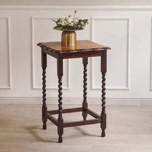 Könnte beinhalten: Ein dunkelbrauner Beistelltisch im antiken Stil mit quadratischer Platte und aufwendig gedrechselten Beinen. Auf dem Tisch steht eine goldfarbene Vase mit weißen Blumen. Der Tisch ist etwa 71 cm hoch.