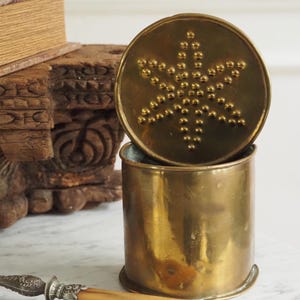 Op de afbeelding: Een vintage messing container met een sterrenhemel deksel. De cilindrische container is open en onthult de binnenkant. Het deksel, met een sterrenpatroon van verhoogde stippen, staat iets open. De container staat op een marmeren oppervlak.