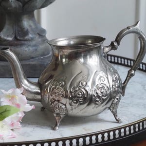 Könnte beinhalten: Eine silberfarbene Teekanne mit geprägten floralen Details. Die Teekanne hat einen gebogenen Ausguss, einen dekorativen Griff und steht auf drei kleinen Füßen. Sie wird auf einem Tisch mit Marmorplatte und dekorativem Rand neben rosa Blüten präsentiert.