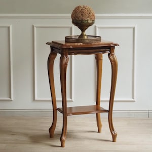 Op de afbeelding: Een hoge houten bijzettafel met vier gebogen poten en een onderste plank. Het tafelblad heeft een rechthoekige marmeren inleg. Op de tafel staat een rond metalen dienblad met een koperen schaal gevuld met een beige, bolvormig object.