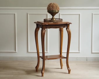 May include: A tall wooden side table with four curved legs and a lower shelf. The tabletop features a rectangular marble inlay. Resting on the table is a round metal tray holding a brass pedestal bowl filled with a textured, spherical beige object.