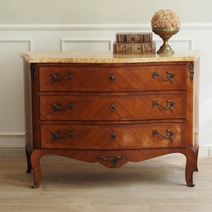 Peut inclure: Une commode en bois avec un plateau en marbre clair, trois tiroirs horizontaux en placage de bois à motif chevron, des poignées en métal bronze et des pieds galbés ornés d'éléments décoratifs en métal sur la base et les coins.