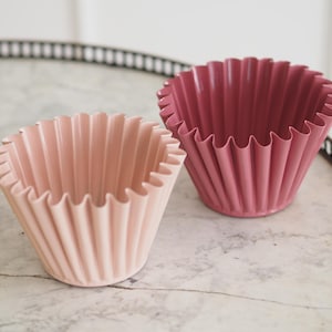 Può includere: Due pirottini scanalati in silicone per cupcake. Uno è rosa chiaro, l'altro rosa scuro. I pirottini sono su un vassoio con motivo marmoreo e bordo in metallo scuro.
