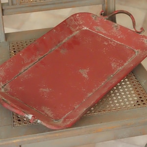 Pode incluir: Uma bandeja de servir retangular de metal com acabamento vermelho desgastado e sinais visíveis de uso. A bandeja possui duas alças de metal curvas nas extremidades opostas, uma borda elevada e uma base plana com textura envelhecida.