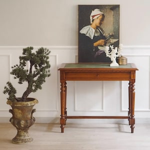 Op de afbeelding: Een antieke houten bureau met een groen leren blad en een lade, versierd met een schilderij en decoratieve items. Een bonsai in pot in een sierlijke urn staat links. De kamer heeft witte lambrisering.