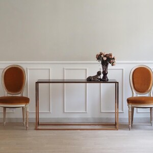 Op de afbeelding: Twee stoelen met een ovale rugleuning, oranje fluwelen bekleding en lichtgekleurde houten frames, geflankeerd door een rechthoekige consoletafel met een donker blad en een metalen frame. Een donkere vaas met droogbloemen en een dierenbeeldje.