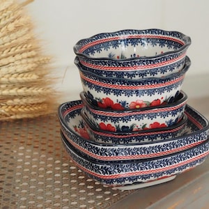 Könnte beinhalten: Ein Stapel aus Keramikschalen und quadratischen Tellern mit cremefarbener Basis, verziert mit komplexen blauen Blumenmustern, roten Blütenakzenten sowie dekorativen rot-blauen geometrischen Rändern an den gewellten und geraden Kanten.