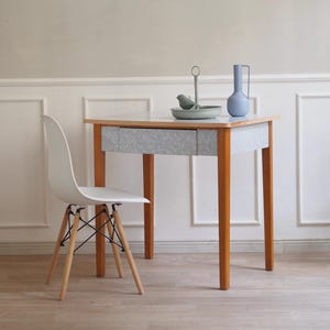 Könnte beinhalten: Ein kleiner Holztisch mit heller Platte und grauer Schublade. Links steht ein weißer Stuhl mit Holzbeinen und schwarzem Metallgestell. Dekorative Gegenstände auf dem Tisch sind eine blaue Vase und ein vogelförmiges Ornament.