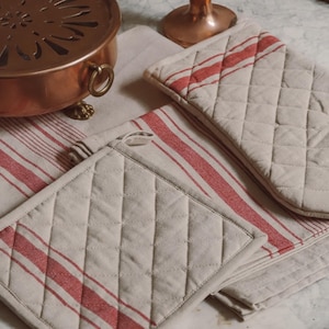Può includere: Un set di tessili da cucina che include un guanto da forno trapuntato e una presina quadrata, entrambi in tessuto beige chiaro con strisce rosse. Gli articoli sono esposti accanto a un contenitore in metallo color rame con coperchio decorativo.