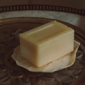 Può includere: Una saponetta rettangolare giallo chiaro poggia su un piccolo piatto a forma di conchiglia bianco sporco. Il portasapone è posto su un vassoio decorativo dorato e ornato, creando un'estetica simile a quella di una spa. Il sapone appare liscio e solido.
