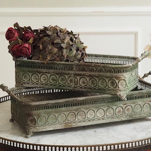Può includere: Due vassoi rettangolari in metallo antico con dettagli decorativi e manici, impilati su un tavolo con piano in marmo. I vassoi hanno una finitura anticata. Un vassoio è riempito di rose bordeaux e verde.