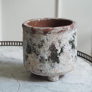 Op de afbeelding: Een cilindervormige terracotta plantenpot met een verweerde, versleten afwerking met vlekken van witte en donkergroene verf over een roodbruine kleibasis. De pot heeft een dikke rand en staat op drie kleine, geïntegreerde voetjes aan de onderkant.