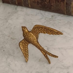 Könnte beinhalten: Ein goldfarbener Serviettenring aus Metall in Form einer fliegenden Schwalbe mit strukturierten Flügeln und einem detaillierten Körper. Der Vogel ist im Flug mit weit ausgebreiteten Flügeln und einem gegabelten Schwanz dargestellt.