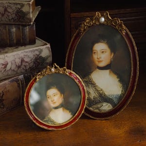 Può includere: Due antiche cornici ovali con ritratti. Una è rotonda, l'altra ovale. Entrambe presentano ritratti di una donna con un choker nero e abiti d'epoca. Le cornici sono rosse e dorate, con dettagli ornati. Libri sullo sfondo.