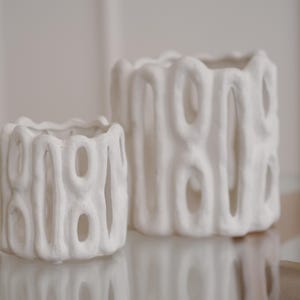 Peut inclure: Deux vases en céramique blanche au design ouvert unique. Les vases présentent une surface texturée avec des découpes ovales, créant un motif intéressant. Le plus petit vase est à gauche d'un plus grand, tous deux sur une surface réfléchissante.