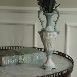 Puede incluir: Un jarrón de cerámica azul claro y blanco con asas, con un diseño de relieve clásico. El jarrón contiene follaje verde y está sobre una mesa con tapa de mármol. Un libro titulado "THEODOR STORM" también está sobre la mesa.