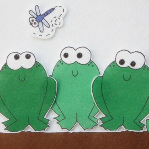 Puede incluir: Tres ranas de fieltro verde con ojos blancos y pupilas negras. Las ranas sonríen y tienen un suelo de fieltro marrón debajo de ellas. Una libélula azul con ojos negros vuela sobre las ranas.