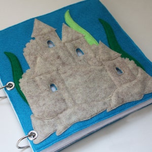 Peut inclure: Un livre en feutre bleu avec un château de sable et des algues. Le livre est relié par des anneaux argentés.