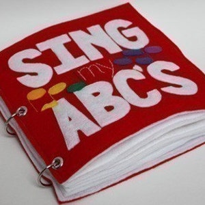 May include: A red felt book with the words "Sing My ABCs" in white felt letters. The book is bound with metal rings and has white felt pages.