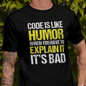 May include: A man wearing a black t-shirt with the text "CODE IS LIKE HUMOR WHEN YOU HAVE TO EXPLAIN IT IT'S BAD" in yellow. He is standing in front of a green leafy wall.