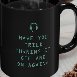 May include: Black coffee mug with white text that reads "Have you tried turning it off and on again?" with a teal headset icon above the text.