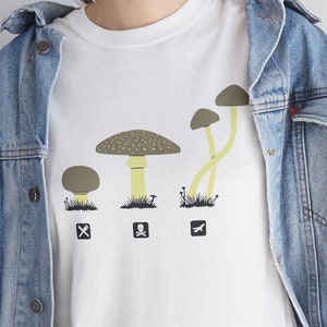May include: White t-shirt with a graphic of four mushrooms in different sizes. The mushrooms are brown and yellow with white spots. The text "Shroom Queens" is printed on the shirt.