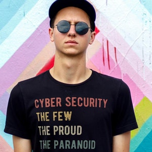 May include: A person wearing a black t-shirt with the text "Cyber Security The Few The Proud The Paranoid" in a vintage style font. The person is wearing a black baseball cap and round sunglasses. The background is a colorful abstract wall with pink, blue, and green stripes.