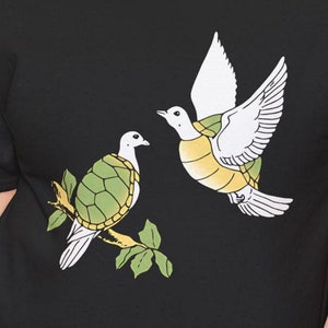 May include: Black t-shirt with a graphic of two white doves with green turtle shells. The doves are perched on a branch with green leaves.