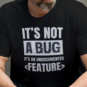 May include: Black t-shirt with the text "IT'S NOT A BUG, IT'S AN UNDOCUMENTED FEATURE" in white. The shirt is worn by a person sitting outside. The text is in a bold, sans-serif font.